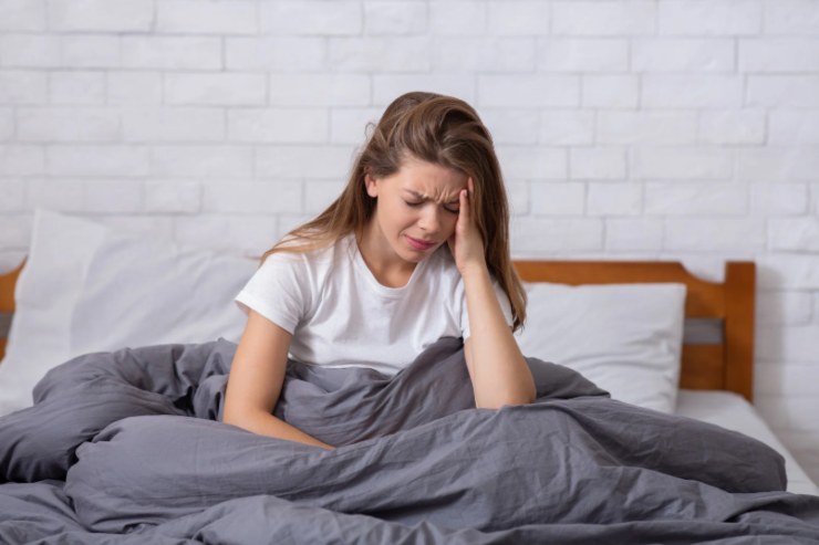 ragazza seduta sul letto sotto le coperte con una mano sulla fronte