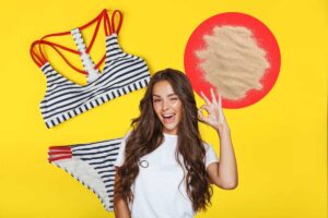 donna che fa ok su sfondo giallo, costume a righe e sabbia in icona